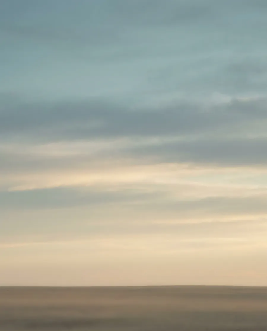 A wide, open prairie sky at golden hour, with soft layered clouds above a flat, distant horizon.